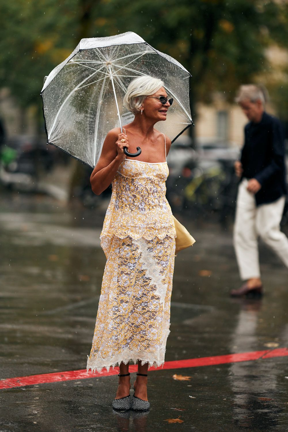 Kobieta w eleganckiej sukience damskiej i okularach przeciwsłonecznych trzyma przezroczystą parasolkę w deszczu.
